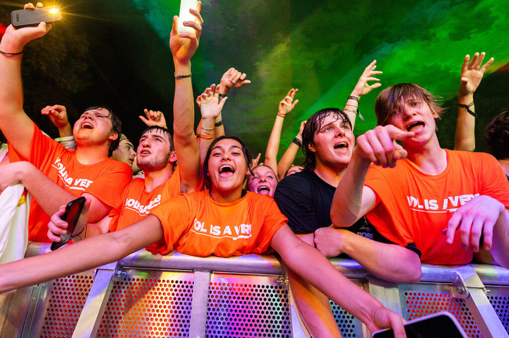 an excited group of students sing along at a concert dressed in orange Vol is a Verb shirts
