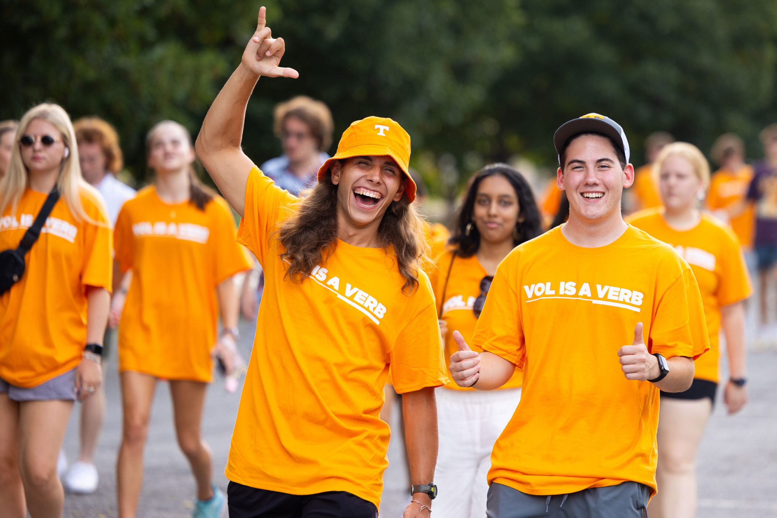 A group of students wearing orange vol is a verb shirts excitedly walk around campus