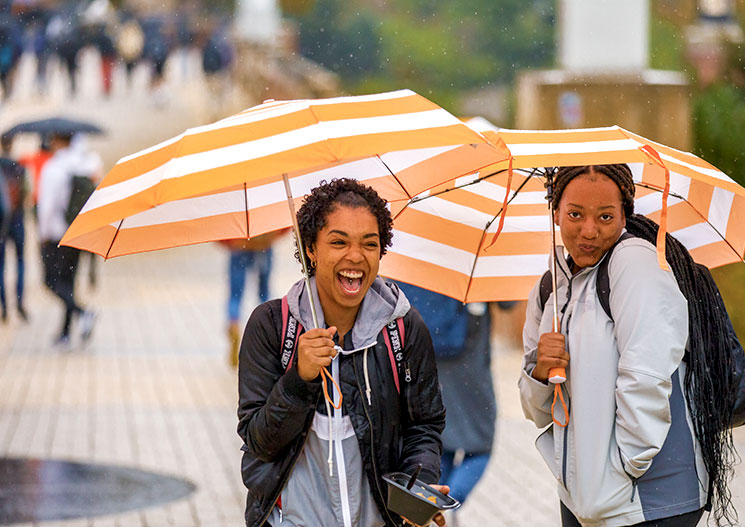 Two students laugh and hold orange and white striped umbrellas as they walk up the ped walkway on a rainy day on campus