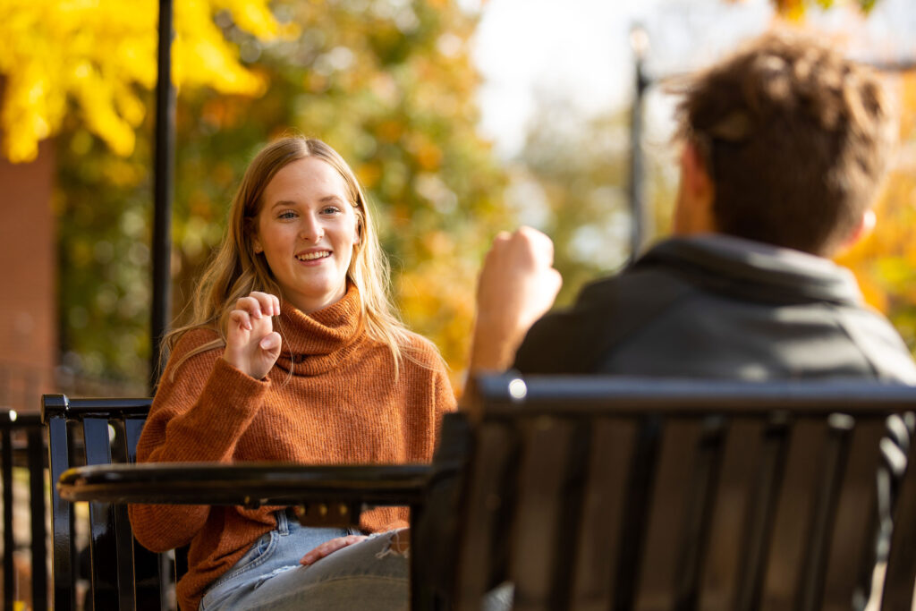 two students sit outside signing to eachother on campus during a beautiful fall day