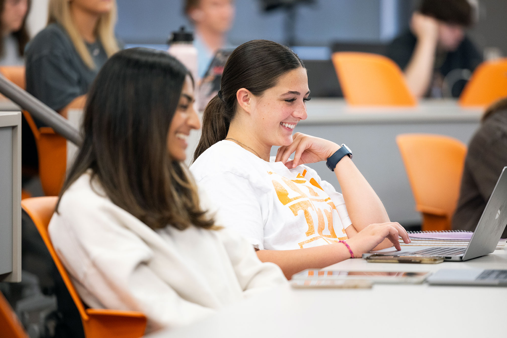 Two women wearing white sweatshirts, sitting in a classroom, smiling and looking at a laptop computer